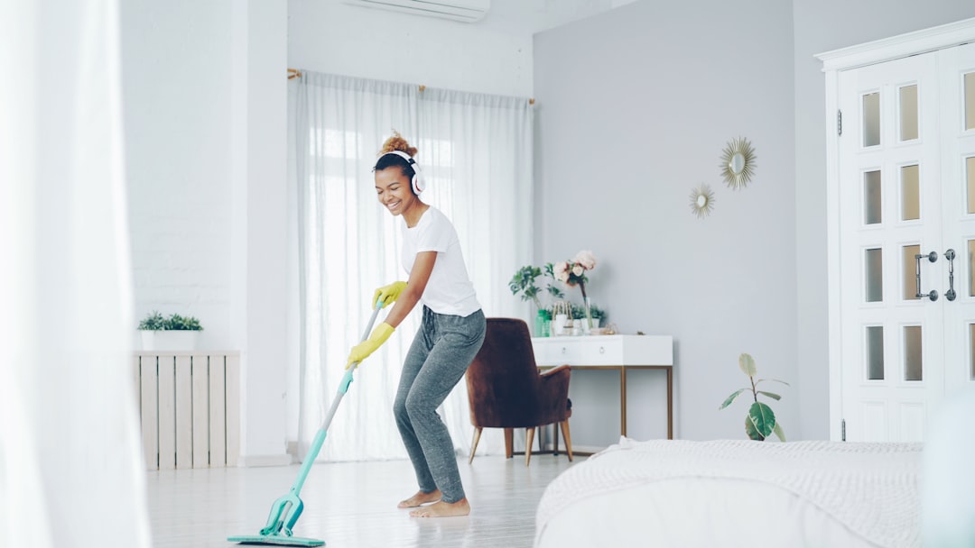 Cheerful African American girl in gloves is mopping floor, listening to music with wireless headphones and dancing enjoying rhythm cleaning her nice modern apartment.
