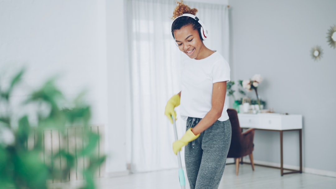 Beautiful African American teenager is mopping floor, dancing and singing listening to radio in wireless headphones. Nice house interior and furniture is visible.